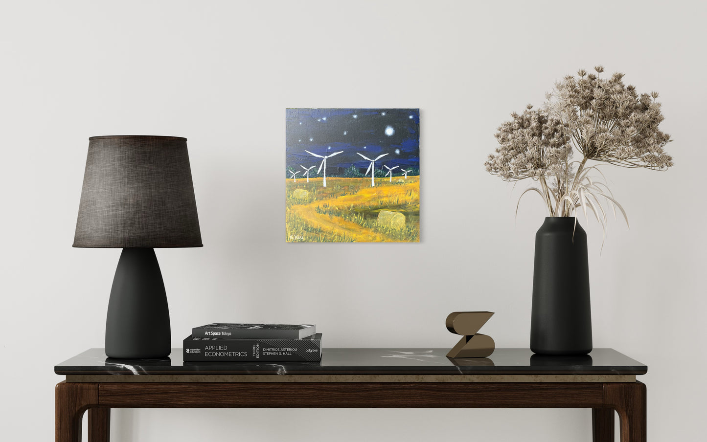 Photo of a painting hung on a white wall of a room with a lamp on the left and vase on the right. They on a table with books and an ornament. The painting is a square acrylic painting on canvas board of a nighttime scene. Dark blue and black sky with white stars and a moon and a yellow field with green grass at either side of a winding path and hay bales. There are six white wind turbines starting from the middle works out to the edges getting smaller.