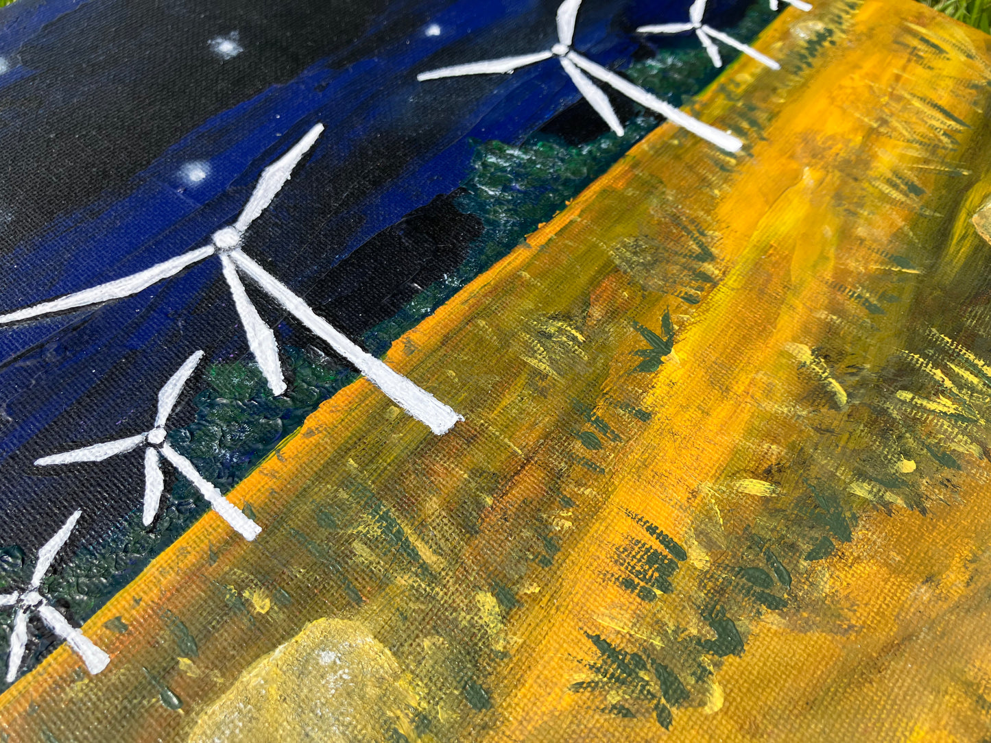 A close up of a square acrylic painting on canvas board of a nighttime scene. Dark blue and black sky with white stars and a moon and a yellow field with green grass at either side of a winding path and hay bales. There are six white wind turbines starting from the middle works out to the edges getting smaller.