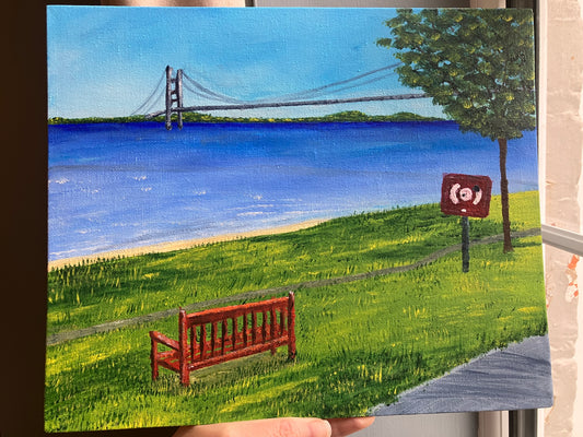 A photo of an acrylic painting on canvas board of the Humber Bridge stands under a light blue sky running from Hessle Foreshore looing over to Grimsby. The blue River Humber flows through the centre of the painting and a slither of light yellow sand can be seen behind the green grass in the forefront of the painting. There is an enpty wooden bench on the grass and a life preserver ring box at the base of a green leafy tree to the right. There's a gray path in the bottom right.