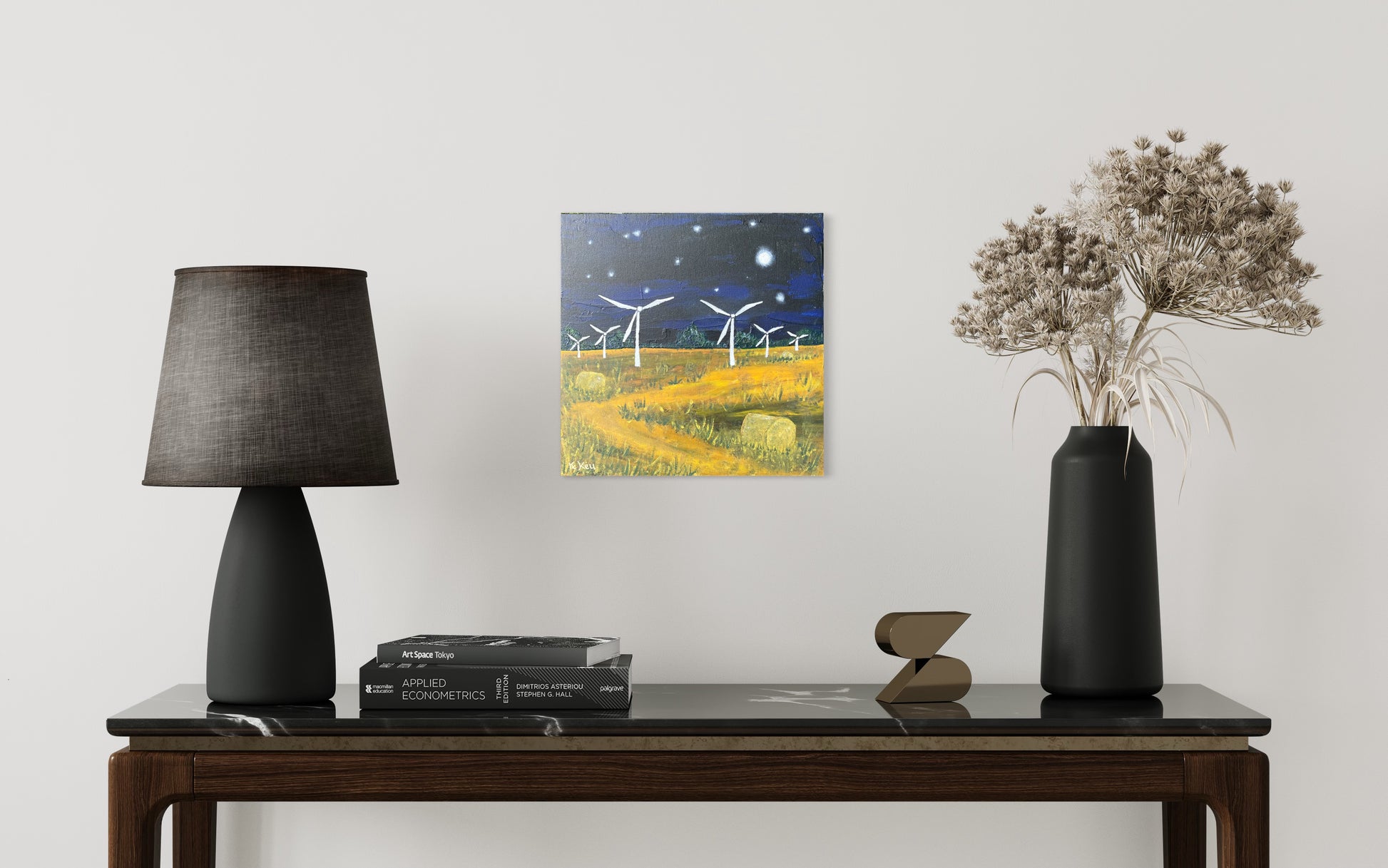 Photo of a painting hung on a white wall of a room with a lamp on the left and vase on the right. They on a table with books and an ornament. The painting is a square acrylic painting on canvas board of a nighttime scene. Dark blue and black sky with white stars and a moon and a yellow field with green grass at either side of a winding path and hay bales. There are six white wind turbines starting from the middle works out to the edges getting smaller.