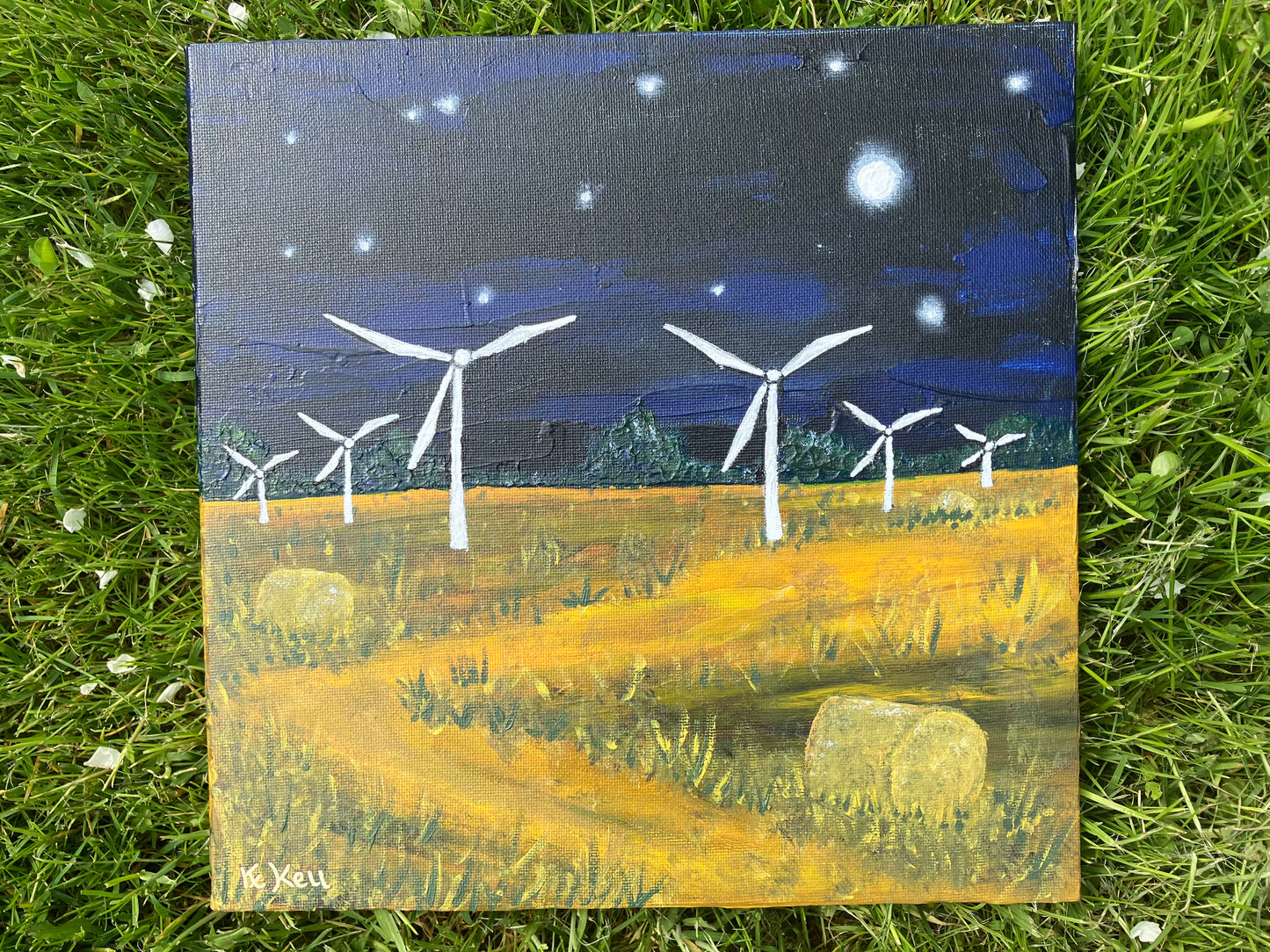 A square acrylic painting on canvas board of a nighttime scene. Dark blue and black sky with white stars and a moon and a yellow field with green grass at either side of a winding path and hay bales. There are six white wind turbines starting from the middle works out to the edges getting smaller.
