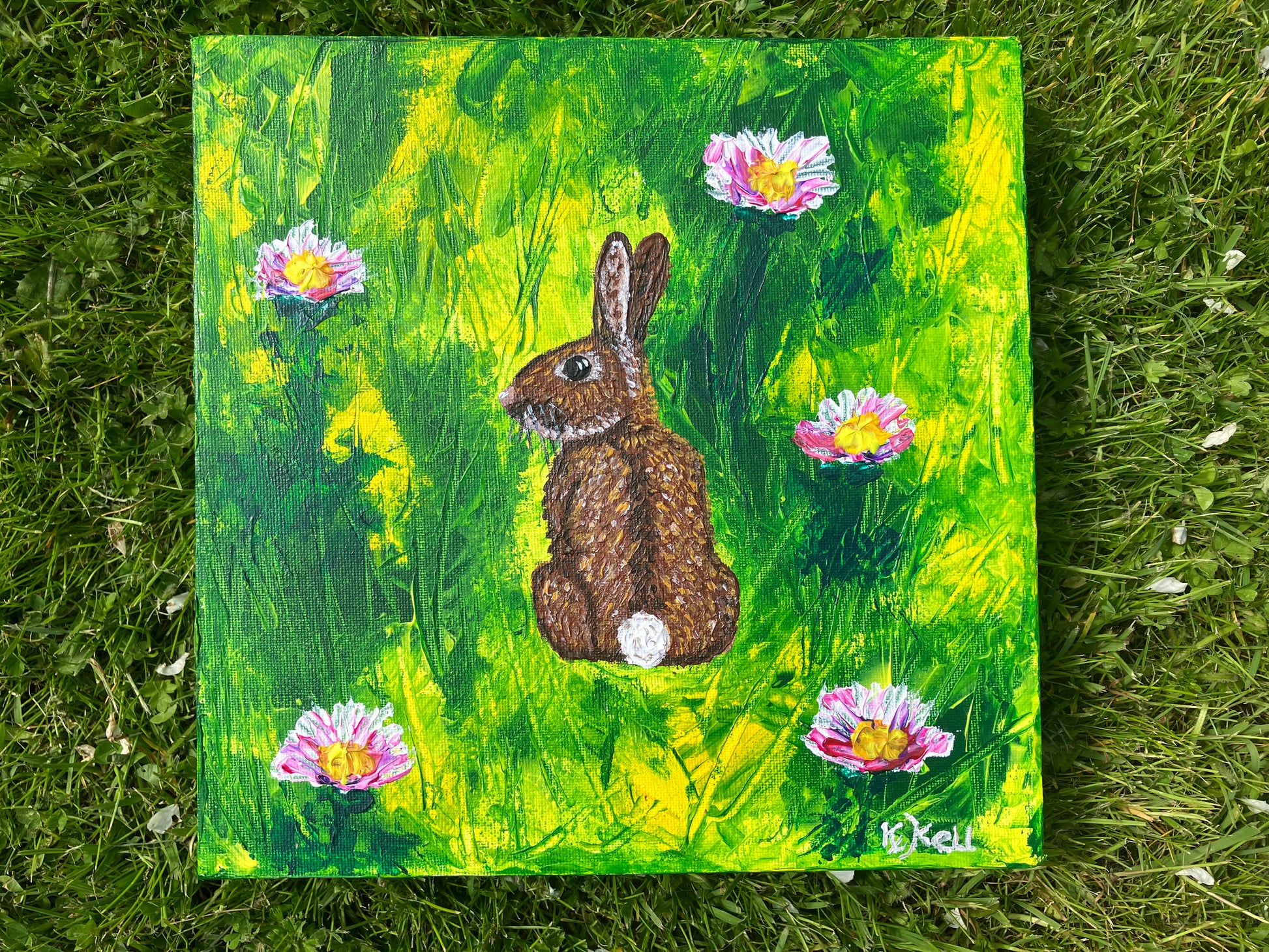 A photo of an acrylic painting on a box canvas of a brown rabbit facing away looking over it's shoulder in a green and yellow field with five white and pink clover flowers. The painting is signed by KtKell in the right hand corner.