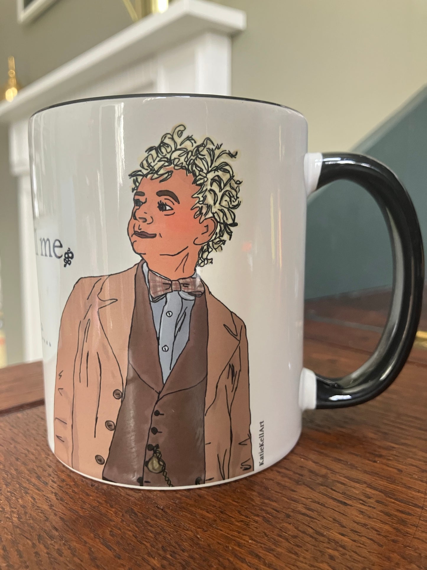 A photo of a white mug with a drawing of Aziraphalefrom Good Omens printed on. The mug has a black handle and rim.