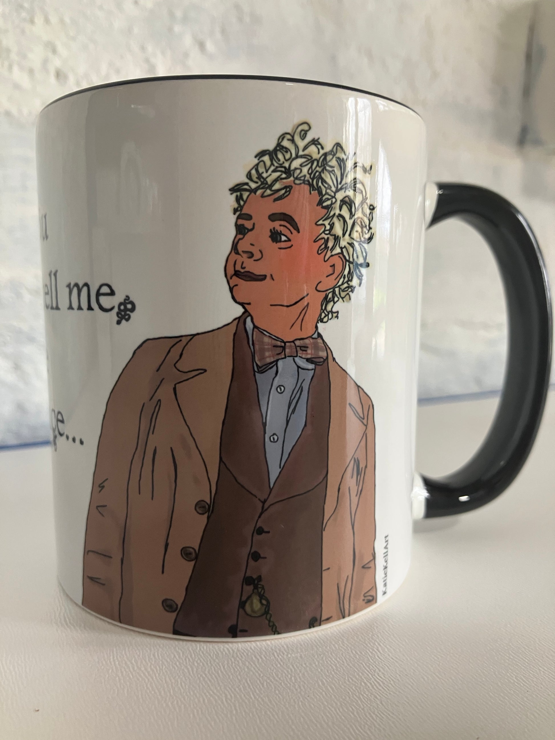 A photo of a white mug with a drawing of Aziraphalefrom Good Omens printed on. The mug has a black handle and rim.