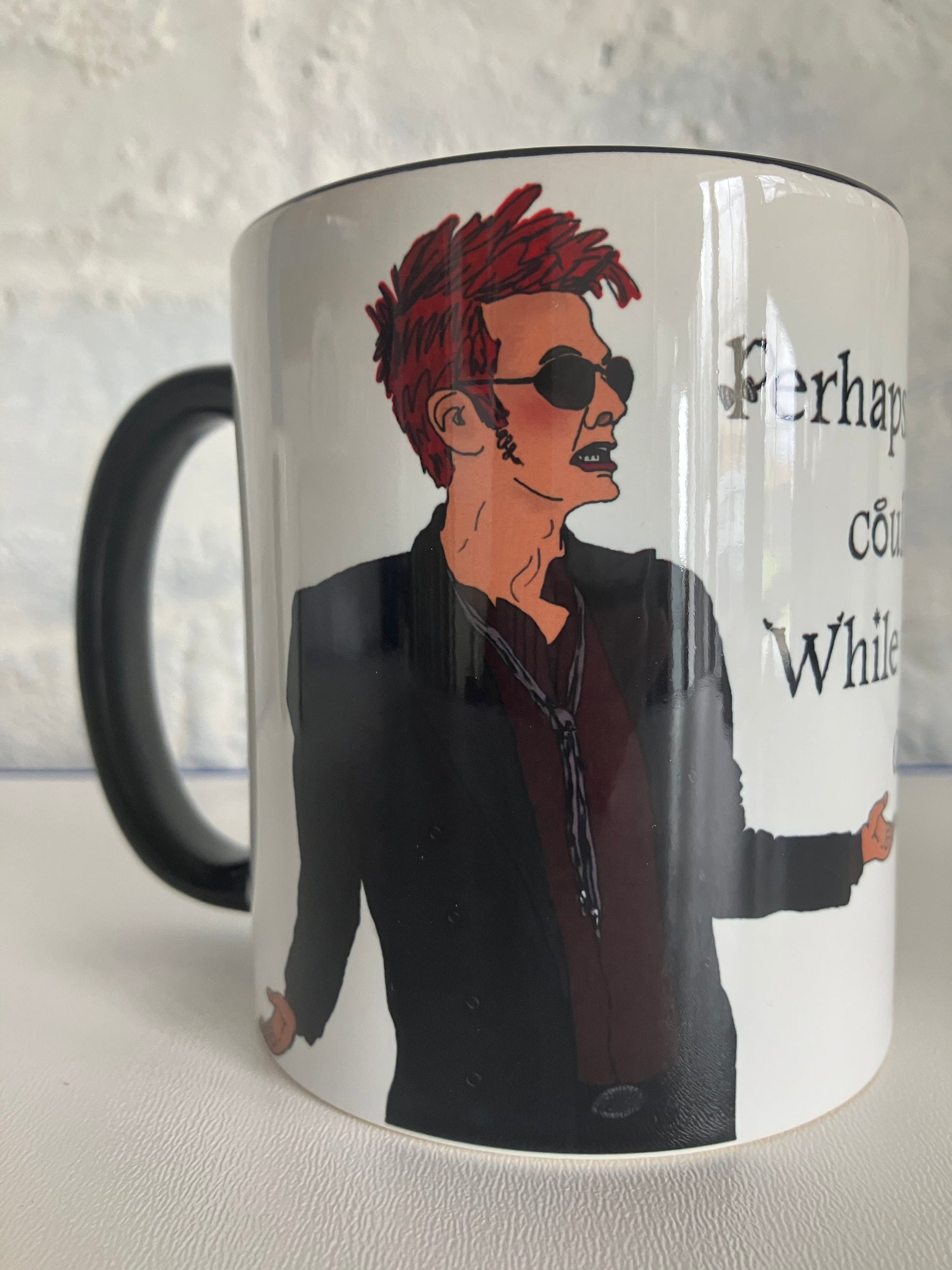A photo of a white mug with a drawing of Crowley from Good Omens printed on. The mug has a black handle and rim.