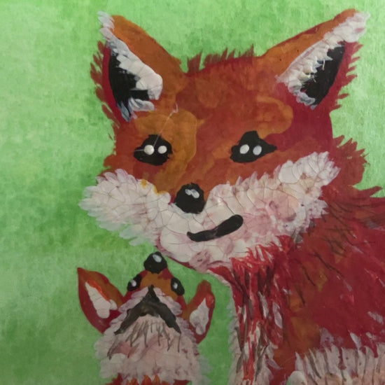 video of a watercolour painting of a red fox and baby with green splash background in a frame on a mantlepiece. 