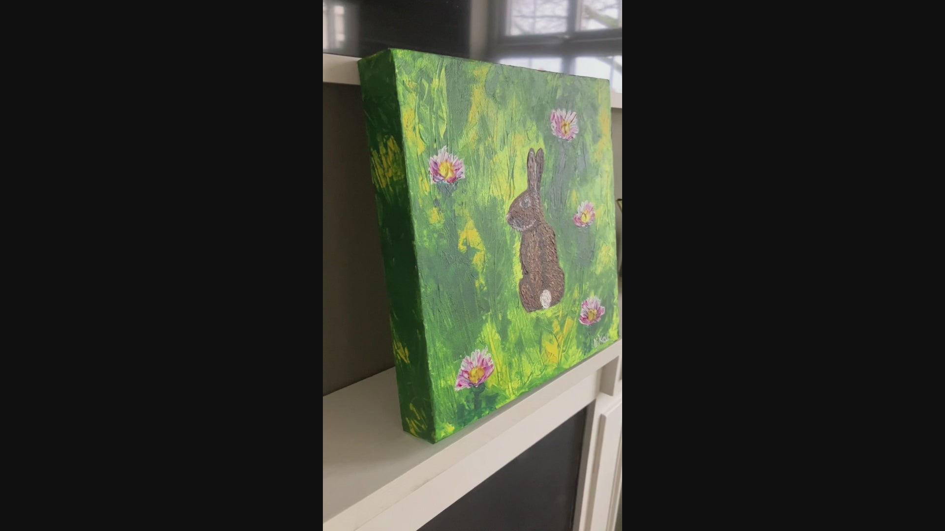A video of an acrylic painting on a box canvas of a brown rabbit facing away looking over it's shoulder in a green and yellow field with five white and pink clover flowers. The painting is signed by KtKell in the right hand corner.