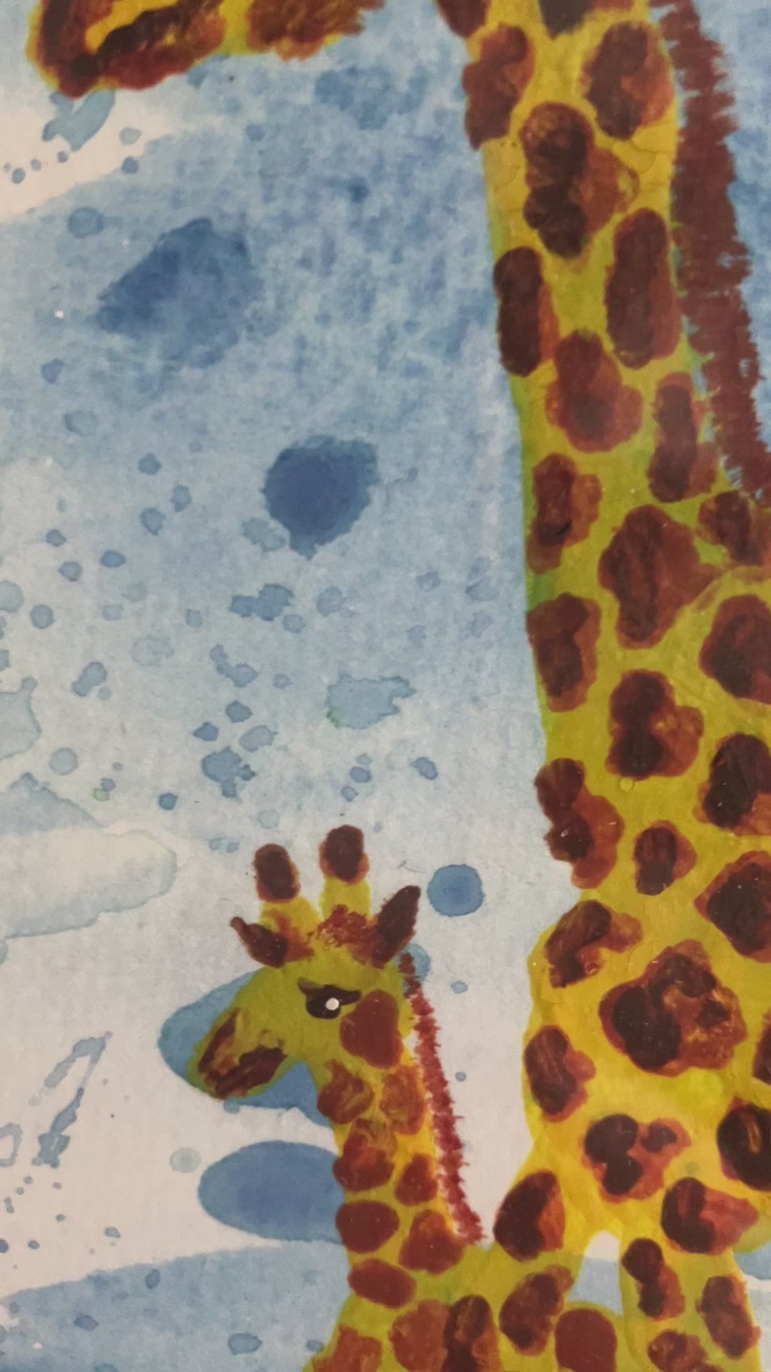 A photo of an original watercolour of a parent giraffe, either mother or father with a baby giraffe looking up at them with a blue splattered background. The painting is in a white frame on a mantlepiece.
