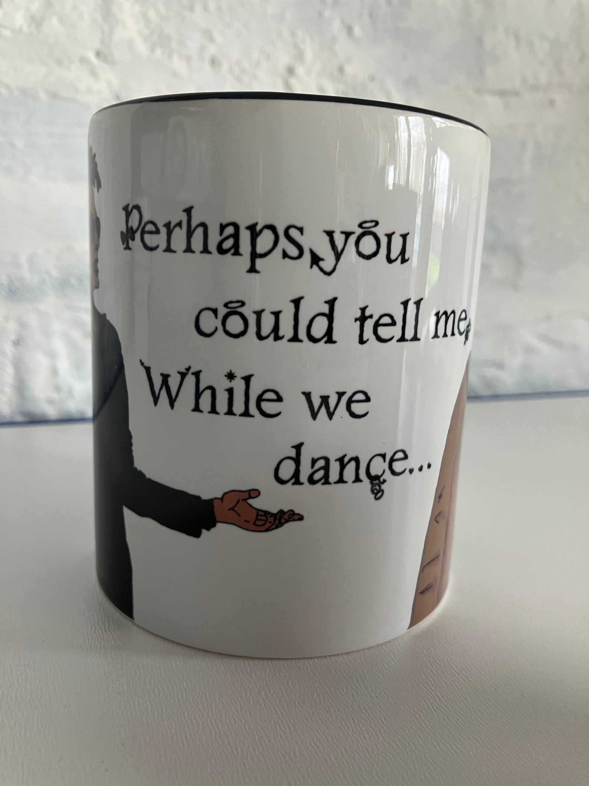 A photo of a white mug with a quote from Good Omens printed on. 'Perhaps you could tell me, while we dance'. The mug has a black handle and rim.