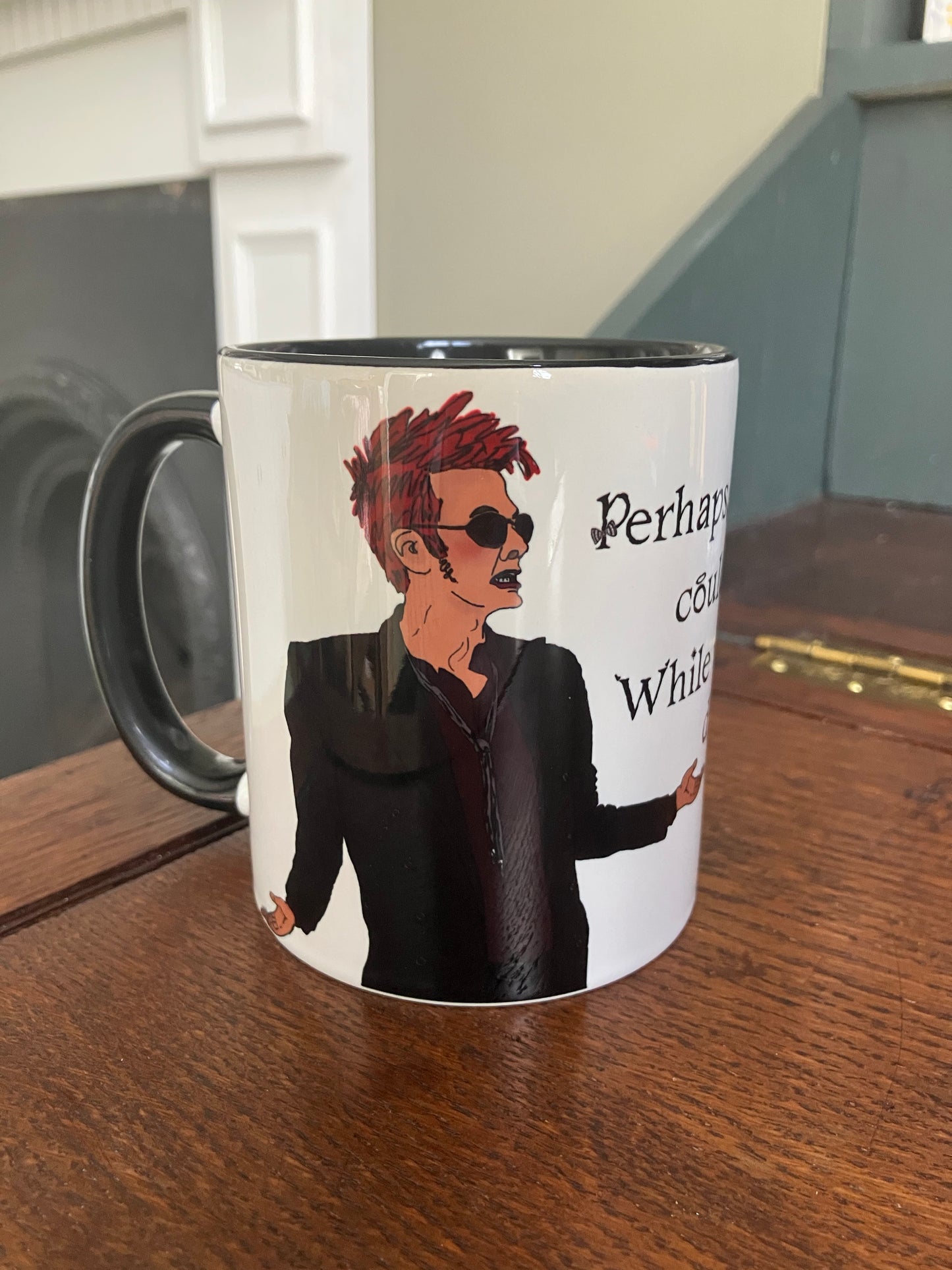 A photo of a white mug with a drawing of Crowley from Good Omens printed on. The mug has a black handle and rim.